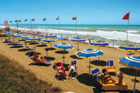 Strand bij glamping met blauw-gele parasols, ligbedden en mensen die genieten van het zonnige weer.