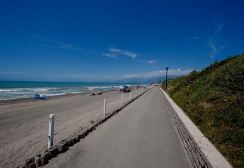 Wide beachfront path next to a mobile home area, sandy beach, a few people, and clear blue sky.