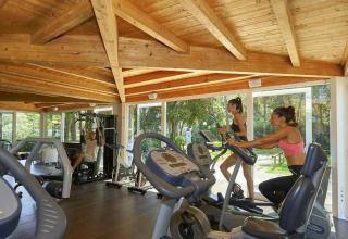 Mensen sporten in een fitnessruimte met houten plafond en grote ramen in een stacaravan omgeving.