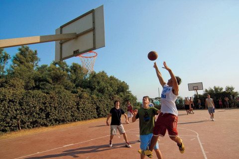 Tieners spelen basketbal op een buitenveld bij een glampingverblijf, omringd door bomen en zonlicht.