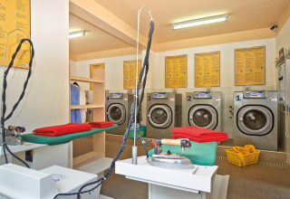 Zelfbedieningswasserette in een stacaravan, met wasmachines, strijktafels en opgevouwen rode handdoeken.