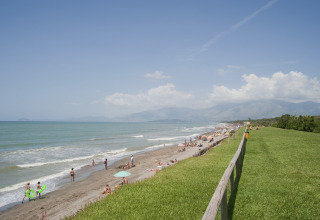 Uitzicht op het strand met badgasten, grasveld en bergen vanaf een staanplaats voor een stacaravan.