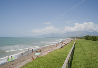 View of a beach with people sunbathing, grassy area, wooden fence, and mountains seen from a mobile home.