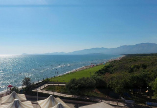 Uitzicht vanuit een stacaravan op een zonnig strand, sprankelende zee, bergen en groene omgeving.