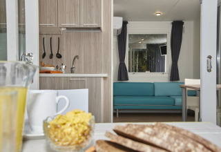 Binnenkant van glamping accommodatie met eettafel, keuken en een turquoise bank bij het raam.