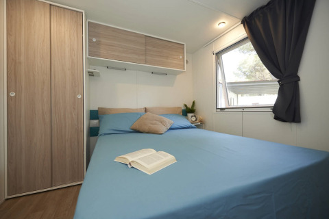 Modern glamping-slaapkamer met blauw bed, houten kastjes, raam en een open boek op het bed.