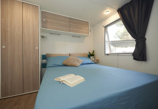 Modern glamping-slaapkamer met blauw bed, houten kastjes, raam en een open boek op het bed.
