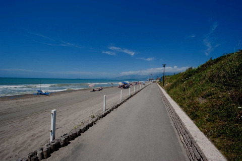 Zonnig strand met een geasfalteerd pad, grasheuvel en mensen die ontspannen op het zand onder parasols.