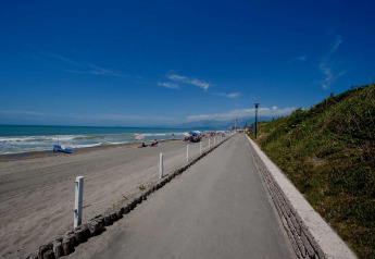 A sunny beach scene with a paved path, grassy slope, and people relaxing under umbrellas on the sand.