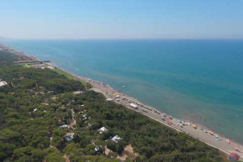 Luchtfoto van een glamping aan het strand met veel groen, parasols en een helderblauwe zee.