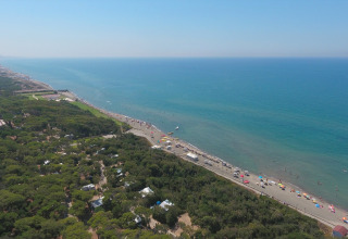 Luchtfoto van een glamping aan het strand met veel groen, parasols en een helderblauwe zee.