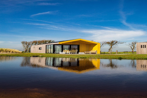 Modern lodge with a yellow roof by a lakeside, reflecting in the water under a clear blue sky.
