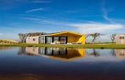 Lodge moderno con techo amarillo junto al lago, reflejado en el agua bajo un cielo azul claro y soleado.