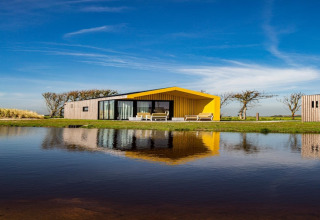 Modern lodge with a yellow roof by a lakeside, reflecting in the water under a clear blue sky.