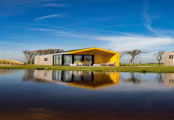 Lodge moderne au toit jaune au bord d’un lac, se reflétant dans l’eau sous un ciel bleu dégagé.