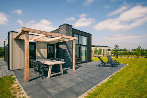 Modern lodge at Holiday Park Zeedijk 2 with patio, dining table, loungers, and green lawn on a sunny day.