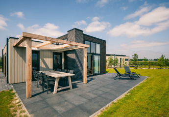 Modern lodge at Holiday Park Zeedijk 2 with patio, dining table, loungers, and green lawn on a sunny day.