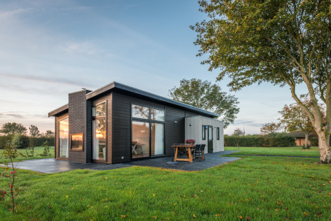 Moderne lodge i Holiday Park Zeedijk 1, omgivet af grønne marker og træer under en klar himmel.