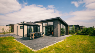 Moderne lodge ved Holiday Park Zeedijk 3 med terrasse, havemøbler og store vinduer på en solrig dag.