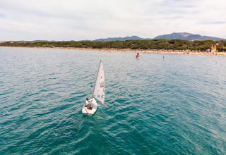 Un velero navega por aguas azules cerca de una playa de arena y colinas verdes en Italia.