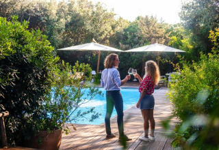 Two people enjoy wine by the poolside, surrounded by lush greenery at Riva degli Etruschi resort in Italy.