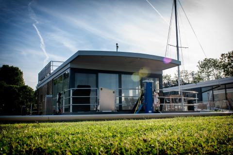 Houseboat moderna chiamata Elysium con terrazza vetrata, ormeggiata su una sponda verde nei Paesi Bassi.