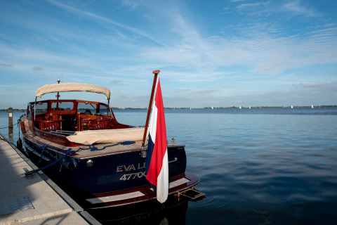 Houseboat 'Elysium' ormeggiata su un lago calmo nei Paesi Bassi con una bandiera e cielo sereno.