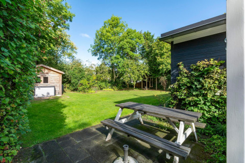 Havebillede fra lodge Eureka 109 i Holland med træbænk, grøn græsplæne, skur og træer i baggrunden.