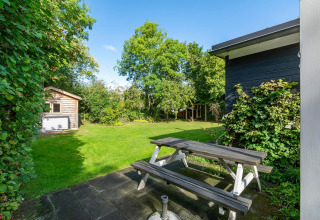 Havebillede fra lodge Eureka 109 i Holland med træbænk, grøn græsplæne, skur og træer i baggrunden.