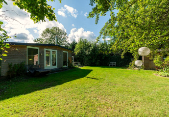 Jardin verdoyant avec lodge en bois Eureka 109 aux Pays-Bas, lanternes suspendues et bancs blancs.