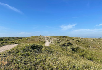 Aussicht auf einen grasbewachsenen Hügel mit Treppe und Bank unter blauem Himmel bei Tipitent de Jutter, Niederlande.