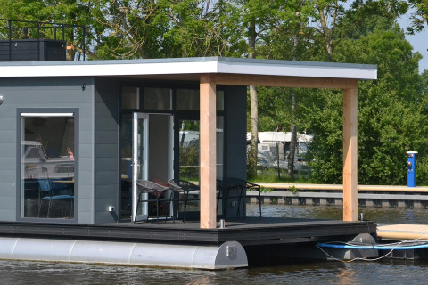 Moderne husbåd «RiggelBrug Sneekermeer» ligger fortøjet på vandet i Holland omgivet af grønne træer.