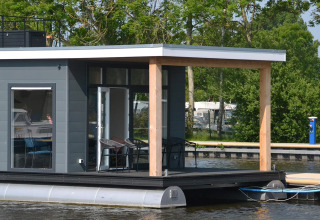 Moderne husbåd «RiggelBrug Sneekermeer» ligger fortøjet på vandet i Holland omgivet af grønne træer.