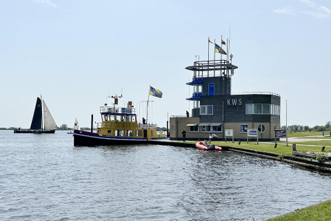 Husbygd båd 'RiggelBrug Sneekermeer' fortøjet ved KWS med sejlbåd på sø og folk på kajen i Holland.
