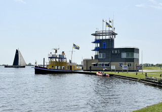 Husbygd båd 'RiggelBrug Sneekermeer' fortøjet ved KWS med sejlbåd på sø og folk på kajen i Holland.