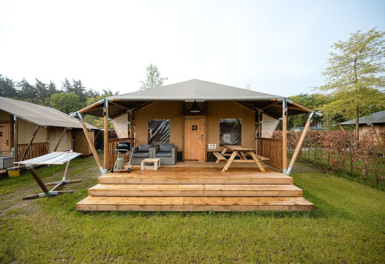 Safari-Zelt Villatent Cottage mit Holzterrasse, Gartenmöbeln und Hängematte auf grünem Gelände.