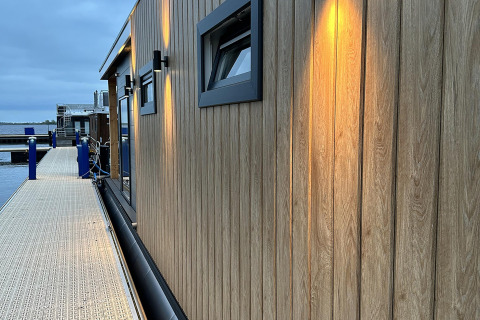 Aftenbillede af en moderne husbåd ved en træbrygge i RiggelBrug Sneekermeer, Holland, med oplyste vægge.