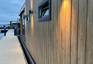Aftenbillede af en moderne husbåd ved en træbrygge i RiggelBrug Sneekermeer, Holland, med oplyste vægge.