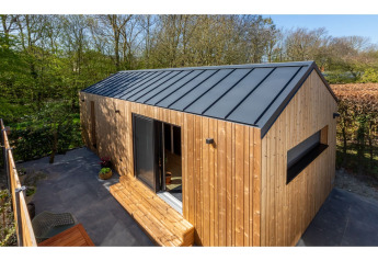 Tiny house en bois avec toit noir, grande porte vitrée et terrasse, nichée dans un jardin verdoyant.