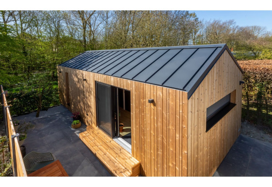 Kleine houten tiny house met zwarte dakbekleding en terras, omgeven door groene natuur en bomen.