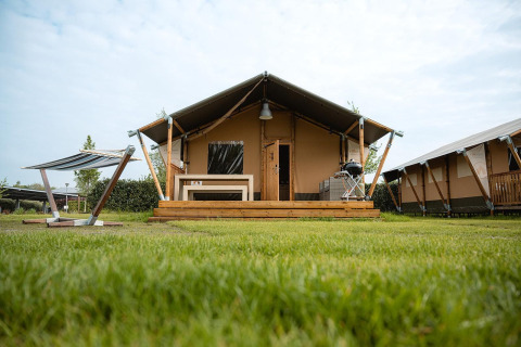 Villatent Nomad safaritent met terras, barbecue en bank op een groen gazon onder een blauwe lucht.