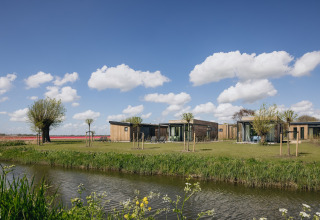 Moderne lodgehytter ved en sø med træer, grøn plæne og blå himmel med skyer i Danmark.