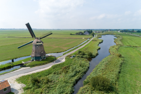 Billede af en traditionel hollandsk vindmølle ved en flod med frodige grønne marker og en blå himmel.