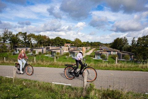 To personer cykler på en vej foran moderne lodge-hytter omgivet af grønne områder og træer.
