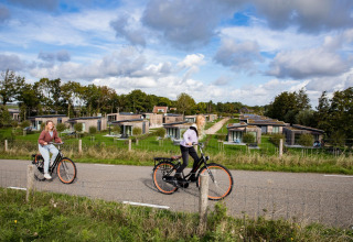 To personer cykler på en vej foran moderne lodge-hytter omgivet af grønne områder og træer.