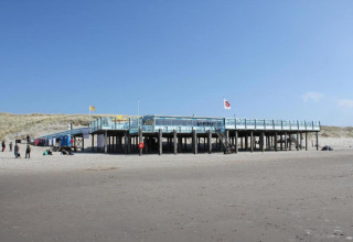 Et strandlodge på pæle med personer på sandet foran, under en klar blå himmel og klitter bagved.