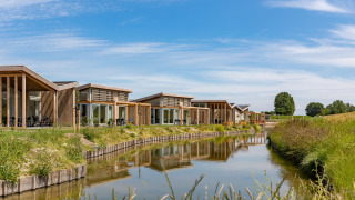 Modern wooden lodges at Water Village 2, lining a peaceful canal with grassy banks and blue sky above.