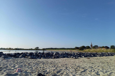 Strandbillede fra Park Wijdenes 3 i Holland med sand, sten, hav og kirketårn i baggrunden.