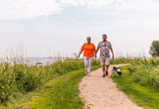 Paar spaziert mit Hund am Wasser entlang, Villa Holiday Park IJsselhof 12, Niederlande, an einem bewölkten Tag.