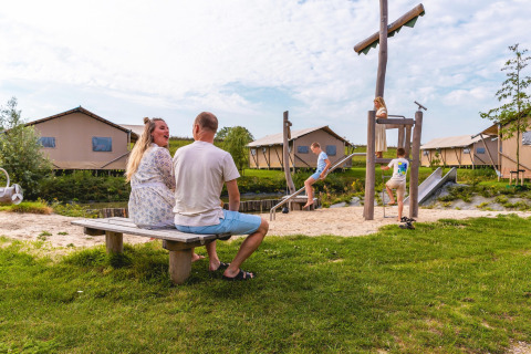 Family relaxing by playground in front of safari tents at Villa Holiday Park IJsselhof, Netherlands.